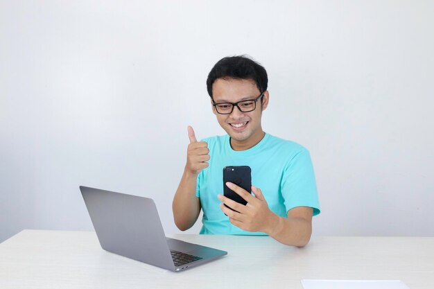 Visage heureux d'un jeune homme asiatique lors d'un appel vidéo ou quoi au téléphone avec un ordinateur portable à côté d'un fond gris isolé Homme indonésien portant une chemise bleue