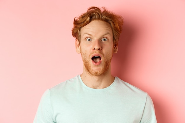 Visage d'un homme rousse surpris réagissant à l'offre promotionnelle cool, haussant les sourcils et haletant, regardant avec admiration la caméra, debout sur fond rose.