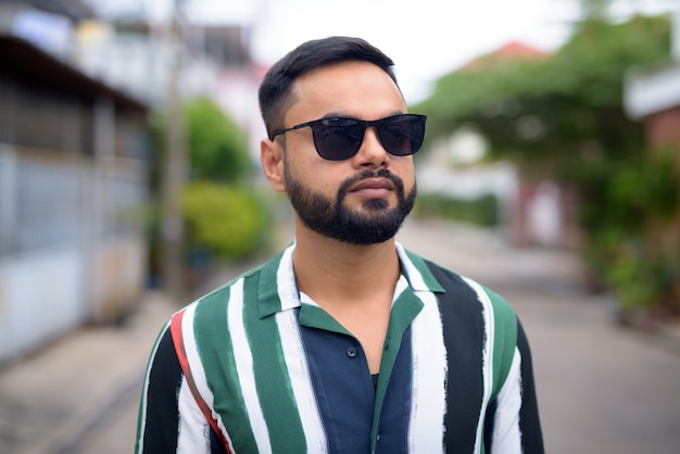 Visage de jeune bel homme indien barbu avec des lunettes de soleil pensant à l'extérieur