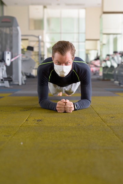 Visage d'un jeune homme avec masque faisant la position de la planche sur le sol au gymnase pendant le coronavirus Covid-19