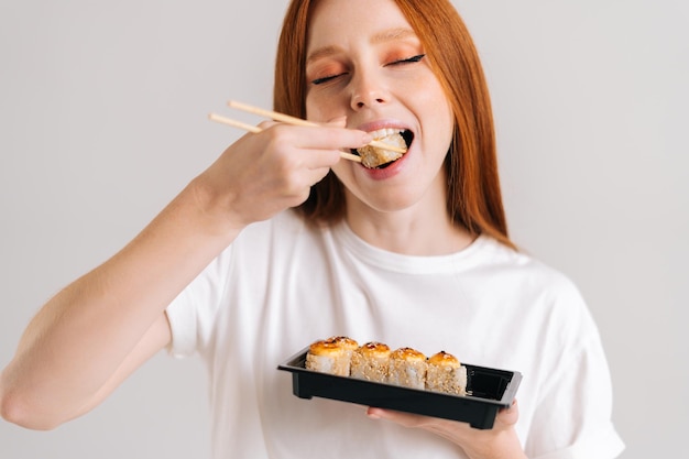 Visage de plan rapproché de jeune femme heureuse avec des yeux fermés mangeant de délicieux rouleaux de sushi avec des baguettes