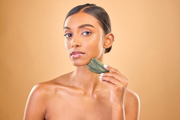 Photo visage portrait soins de la peau et femme avec gua sha en studio isolé sur fond marron massage dermatologique et modèle féminin indien sérieux avec cristal de jade ou pierre pour un traitement de la peau saine