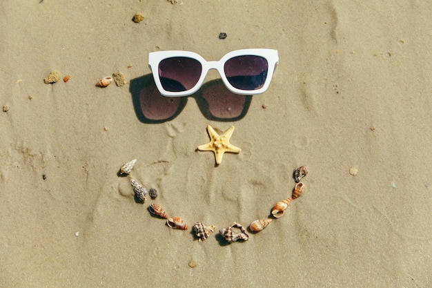 Visage souriant dessiné sur du sable avec des coquillages et des lunettes de soleil