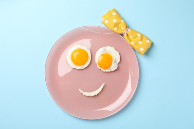 Visage souriant fait de plaque avec des œufs au plat sur fond bleu, vue de dessus