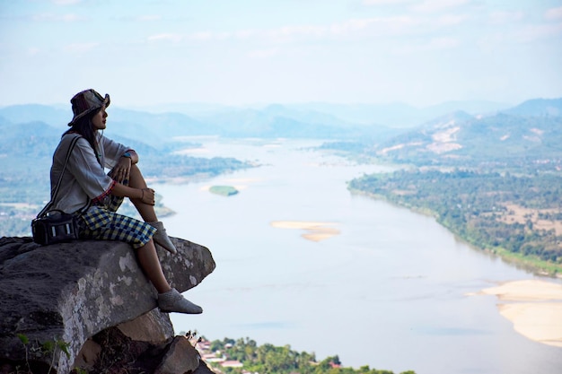 Visite de voyage d'une femme thaïlandaise asiatique et posant assis sur la pierre de crête des falaises du temple Wat Pha Tak Suea avec vue sur le paysage de la ville de Nongkhai et les loas et le fleuve Mékong à Nong Khai en Thaïlande