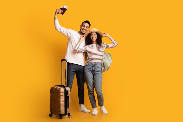Visites en famille. Jeunes époux arabes joyeux avec valise, passeports et billets posant sur fond jaune en studio, couple de touristes du Moyen-Orient prêt pour un voyage de vacances, pleine longueur, espace de copie
