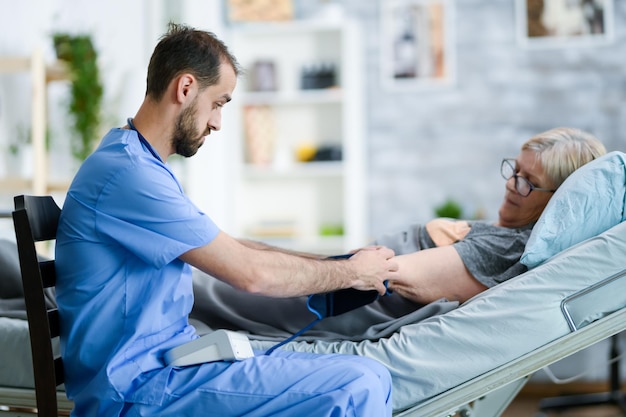 Visiteur de santé barbu dans une maison de retraite avec stéthoscope vérifiant la tension artérielle d'une vieille dame à l'aide d'un appareil numérique.