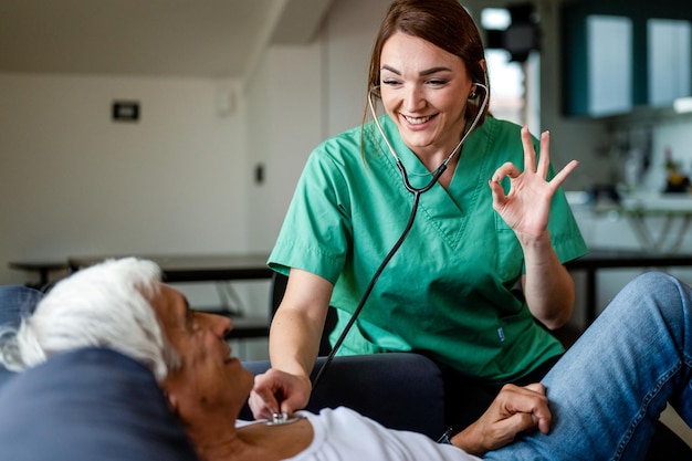 Visiteur de santé utilisant un stéthoscope pour vérifier la santé d'un homme âgé, jeune infirmière faisant des gestes ok avec la main lors d'une visite à domicile, concept de soins de santé à domicile