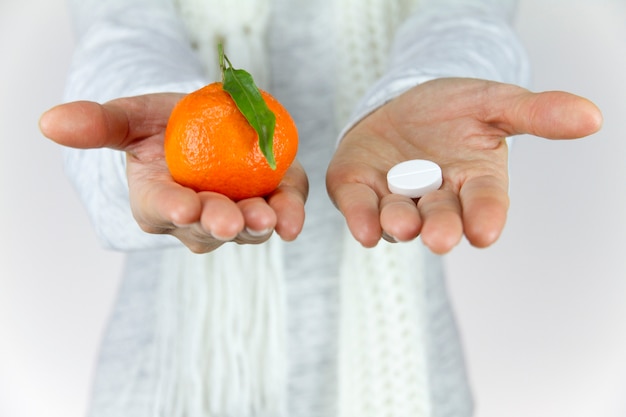 Vitamines de fruits ou de drogues? Une jeune femme malade avec un foulard sur le cou montre un mandarin dans sa main droite et une aspirine sur sa gauche