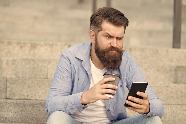 Vivre la vie en ligne Réseaux sociaux Communication en ligne de la vie moderne Mec avec fond d'espace urbain smartphone Bel homme barbu relaxant téléphone portable et tasse à café Technologie moderne