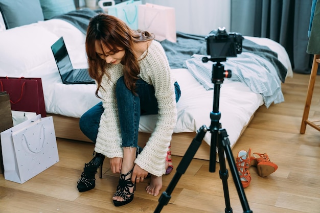 Le vlogger de beauté asiatique se plie et essaie des chaussures à talons noires sur le lit par un appareil photo numérique tout en filmant une vidéo de déballage sur le réseau social dans la chambre à la maison.