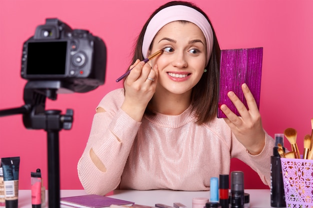 Vlogger souriant heureux est assis devant la caméra et tient la brosse. Jeune femme regarde le miroir et applique le fard à paupières