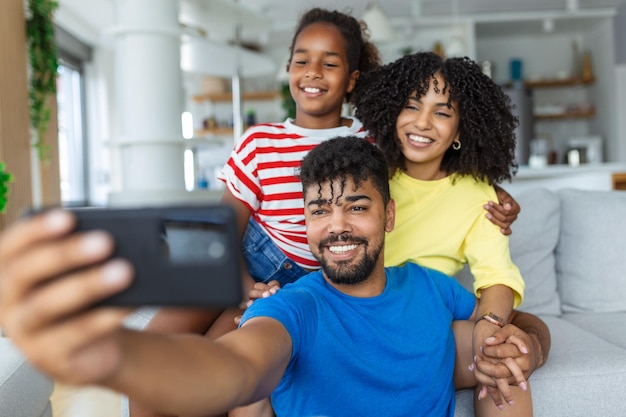 Des vloggers de famille joyeux avec une fille en train de se faire un selfie, d'enregistrer un vlog, de faire un appel vidéo au téléphone, de s'amuser ensemble dans le salon, des parents et une petite fille riant en utilisant un téléphone portable, une application en ligne drôle.