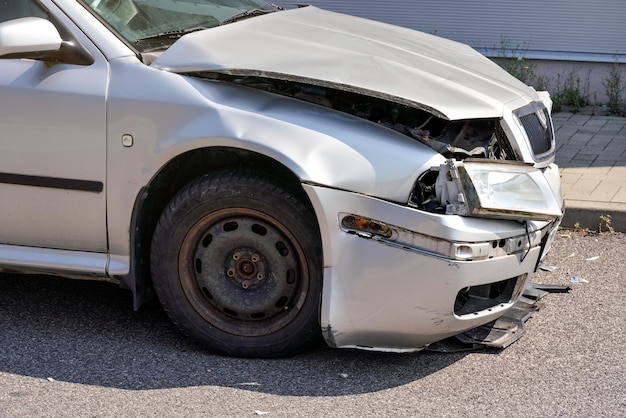 Voiture argentée avec son avant écrasé, plaques bosselées et cassées sur route goudronnée, détail de la partie endommagée