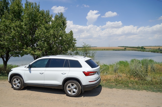 Une voiture blanche se dresse dans la nature près d'un arbre sur fond de lac et de ciel avec des nuages