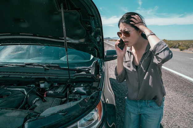 voiture cassée rendant la conductrice folle. une jeune fille asiatique ouvre le moteur en le regardant et se sent confuse. Dame inquiète parlant au téléphone avec un service d'aide d'urgence sur une autoroute vide