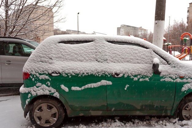 Une Voiture Compacte Verte Sale Dans La Neige Ne Peut Pas Aller. La Première Chute De Neige. Voitures Couvertes De Neige Dans La Ville. Problèmes De Transport, Neige Mal Nettoyée.