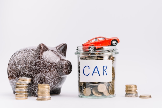 Voiture jouet sur le pot rempli de pièces de monnaie et pile de pièces et tirelire sur fond blanc