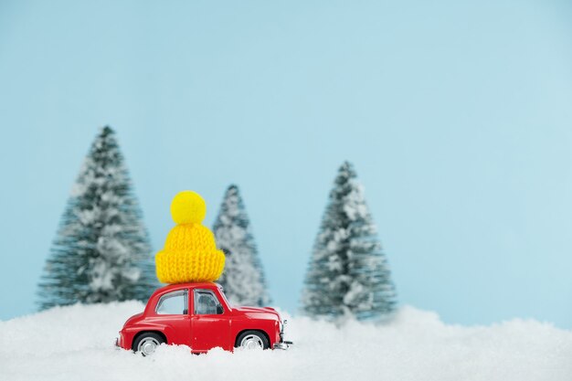 Voiture rouge de Noël avec chapeau jaune tricoté dans une forêt de pins enneigée