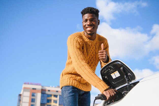 Voitures électriques, concept EV, carburant écologique. Portrait de jeune homme noir souriant, rechargeant sa voiture électrique de luxe moderne