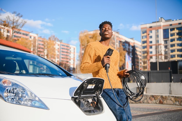 Voitures électriques, concept EV, carburant écologique. Portrait de jeune homme noir souriant, rechargeant sa voiture électrique de luxe moderne