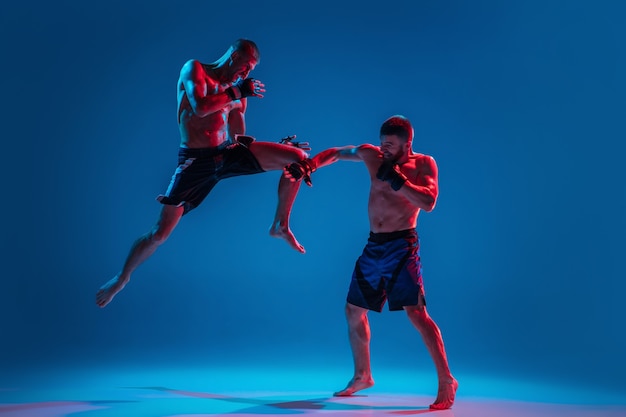 En vol. MMA. Deux combattants professionnels poinçonnant ou boxant isolés sur fond bleu studio en néon. Ajustez les athlètes ou les boxeurs caucasiens musclés qui se battent. Sport, compétition et émotions humaines, annonce.