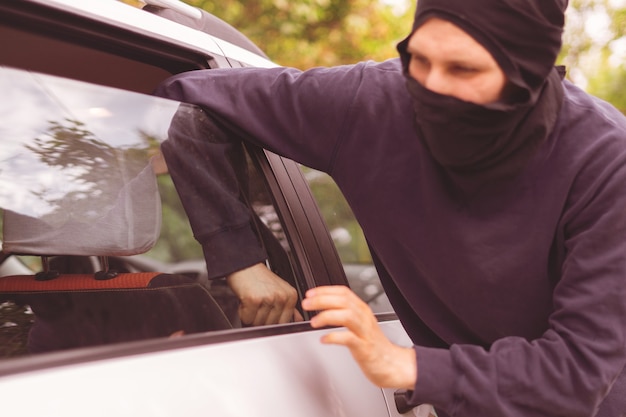 Un voleur masqué déverrouille et ouvre la fenêtre de la voiture pour voler des biens