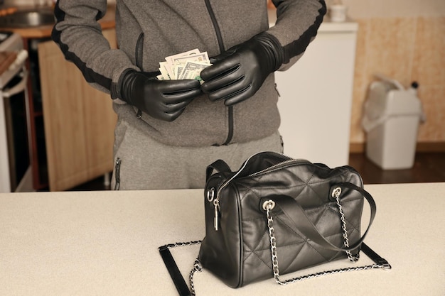 Voleur en tenue noire et gants voir dans le sac de femmes volé ouvert Le voleur sort l'or et l'argent d'un sac à main de femme dans la cuisine