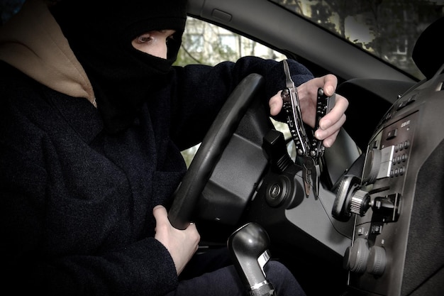 Un voleur de voiture essaie d'ouvrir le contacteur d'allumage avec un outil Le voleur de voiture s'est caché le visage dans une cagoule