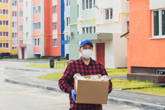 Un volontaire masculin portant un masque de protection marche dans la rue avec une boîte d'épicerie, une œuvre de bienfaisance.