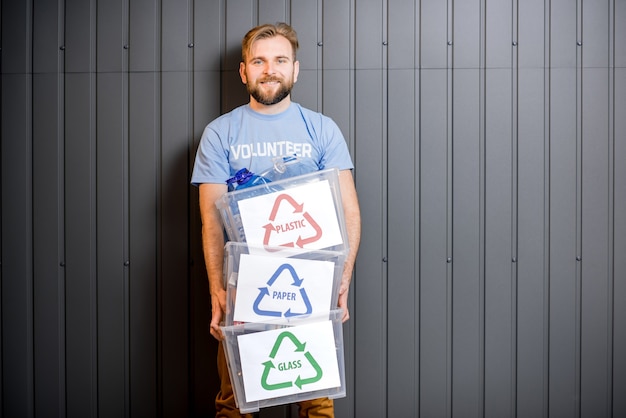 Volontaire masculin en t-shirt bleu tenant des conteneurs avec des déchets triés debout à l'intérieur sur le fond du mur gris