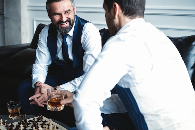 Votre déménagement! Deux jeunes hommes beaux en costumes complets jouant aux échecs et souriant assis à l'intérieur.