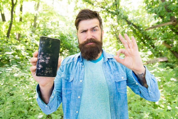 Voudrais souhaiter le meilleur. un homme barbu bouleversé montre un geste ok au téléphone. Hipster mature avec barbe. s'est perdu dans la forêt. homme brutal avec téléphone. style hipster. gars dans la forêt utilise le gps. camping d'été.