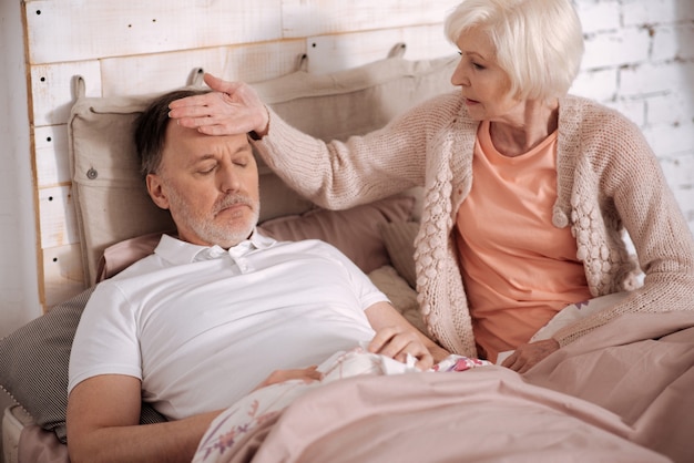 Vous avez froid. Femme âgée assise sur le lit près de son mari malade et sentant sa température corporelle en touchant son front.