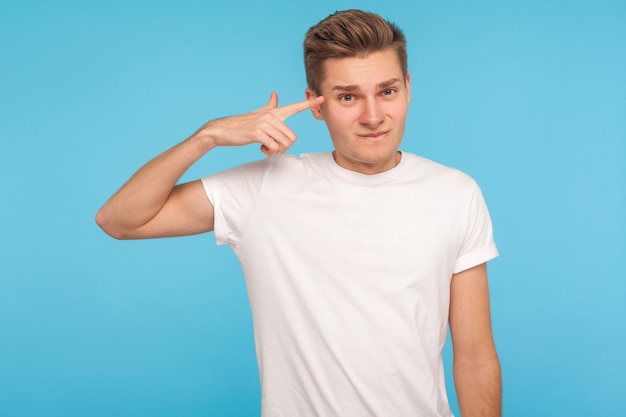 Vous êtes idiot Homme mécontent en t-shirt blanc décontracté tenant le doigt contre sa tempe faisant un geste stupide et fou blâmant dans un discours muet insensé tourné en studio intérieur isolé sur fond bleu