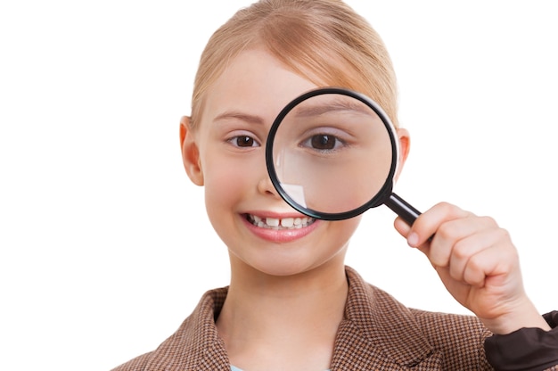 Photo vous examiner. joyeuse petite fille en tenue de soirée vous examinant avec une loupe en se tenant debout isolée sur blanc