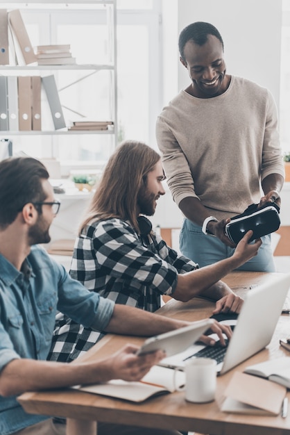 Vous ne regretterez pas ! L'équipe de développement coopère dans son bureau tandis qu'un bel homme aux cheveux longs donne un casque VR à son collègue