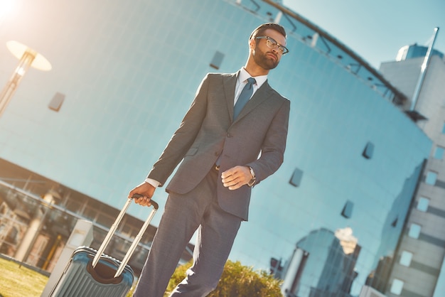 Voyage d'affaires jeune et bel homme barbu en costume complet tirant ses bagages et regardant