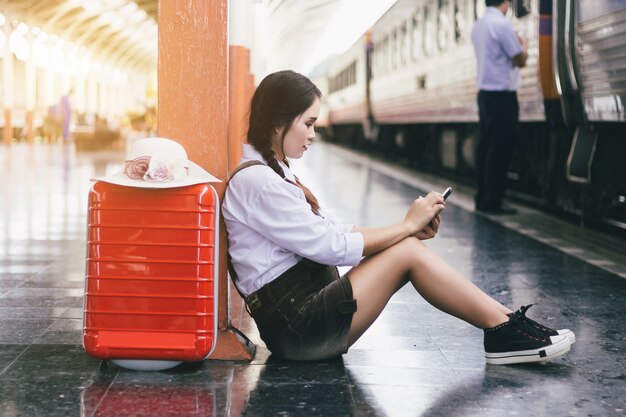 Voyage femme asiatique enceinte regarder sur le smartphone avec une valise rouge à la gare.