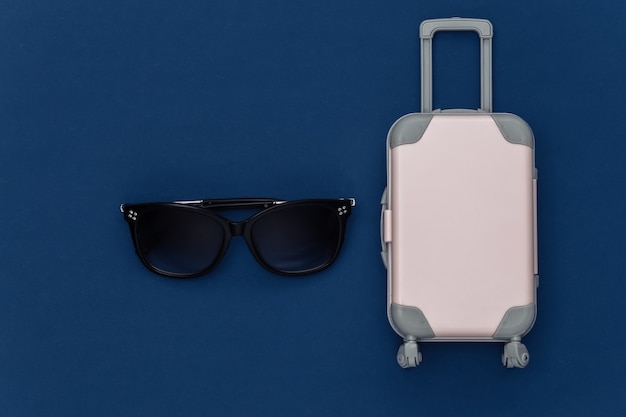 Voyage à plat. Mini valise de voyage en plastique, lunettes de soleil sur fond bleu classique. Style minimaliste. Vue de dessus