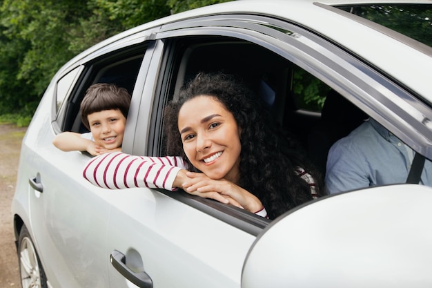 Voyage en voiture. Heureuse jeune famille avec petit fils chevauchant une voiture ensemble, joyeuse mère et enfant de sexe masculin regardant par les fenêtres et souriant à la caméra, profitant de voyages en plein air en automobile, gros plan