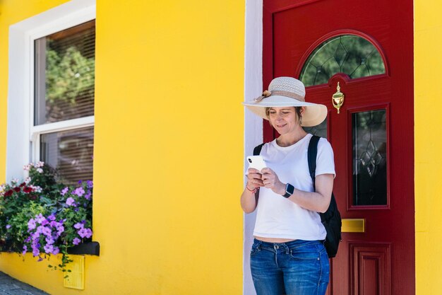 Voyageur blond laissant la maison jaune colorée tout en bavardant au téléphone avec le sac à dos et le chapeau