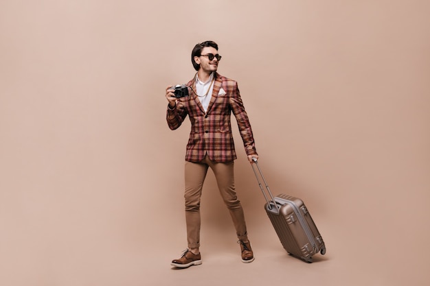 Voyageur élégant en pantalon beige, chaussures en cuir marron, veste à carreaux, tenant un appareil photo et une valise, portant des lunettes de soleil, posant contre un mur de pêche et regardant de côté