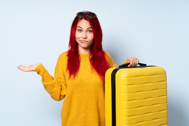 Voyageur femme tenant une valise isolée sur le mur bleu ayant des doutes tout en levant les mains