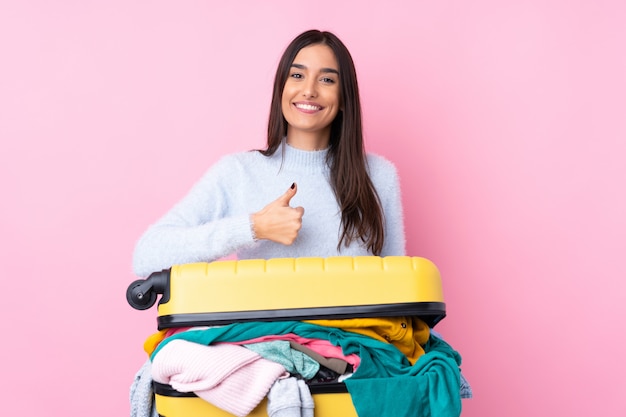 Voyageur femme avec une valise pleine de vêtements sur un mur rose isolé donnant un coup de pouce geste