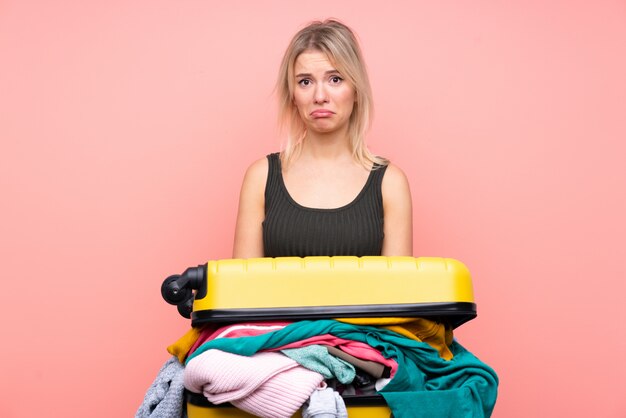 Voyageur femme avec une valise pleine de vêtements sur mur rose isolé triste