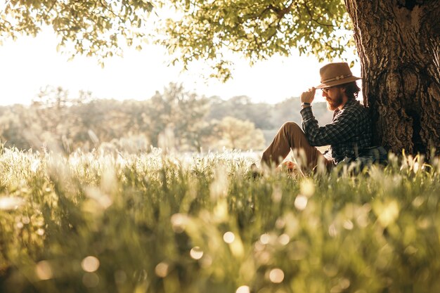 Voyageur hipster réfléchi assis sous un arbre sur un pré vert