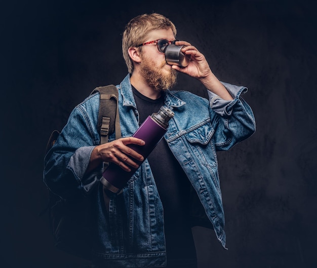 Un voyageur hipster rousse vêtu d'une veste en jean tient un thermos et boit un thé sur fond sombre.