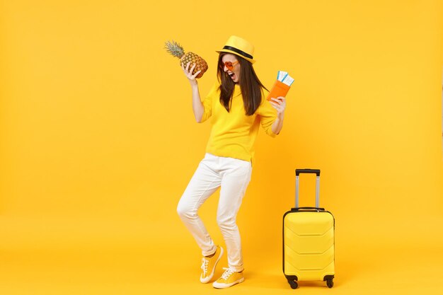 Voyageur hurlant touriste femme au chapeau tenant des billets de passeport fruits d'ananas frais isolés sur fond orange jaune. Passager voyageant à l'étranger en week-end. Concept de voyage de vol aérien.