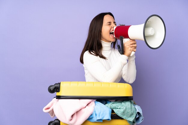 Voyageur jeune femme avec une valise pleine de vêtements sur le mur violet criant à travers un mégaphone